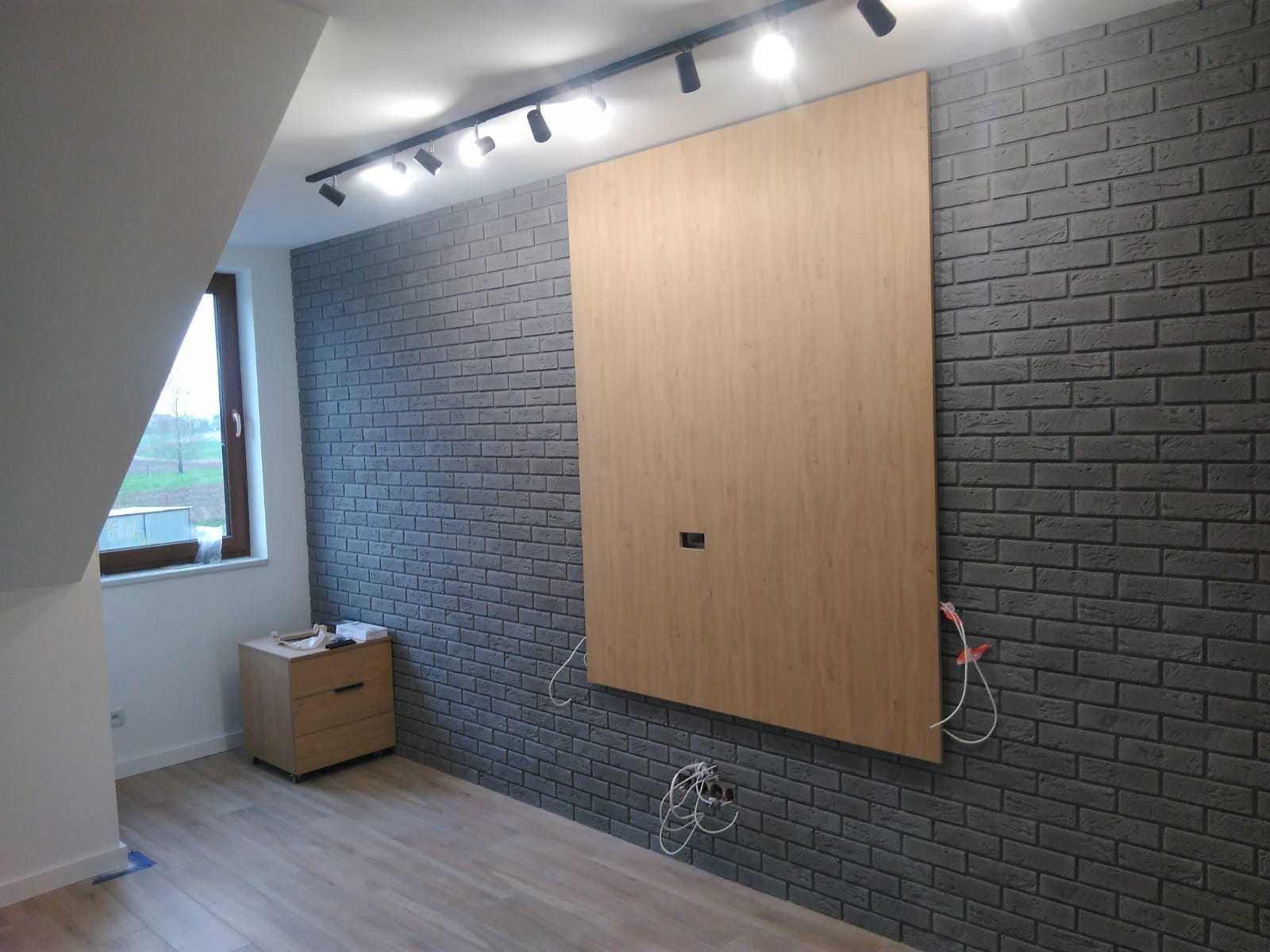Pokój z szarą ścianą z cegły, zamontowanym panelem z jasnego drewna z otworem na kable, oświetleniem sufitowym i drewnianą podłogą w skosach poddasza.