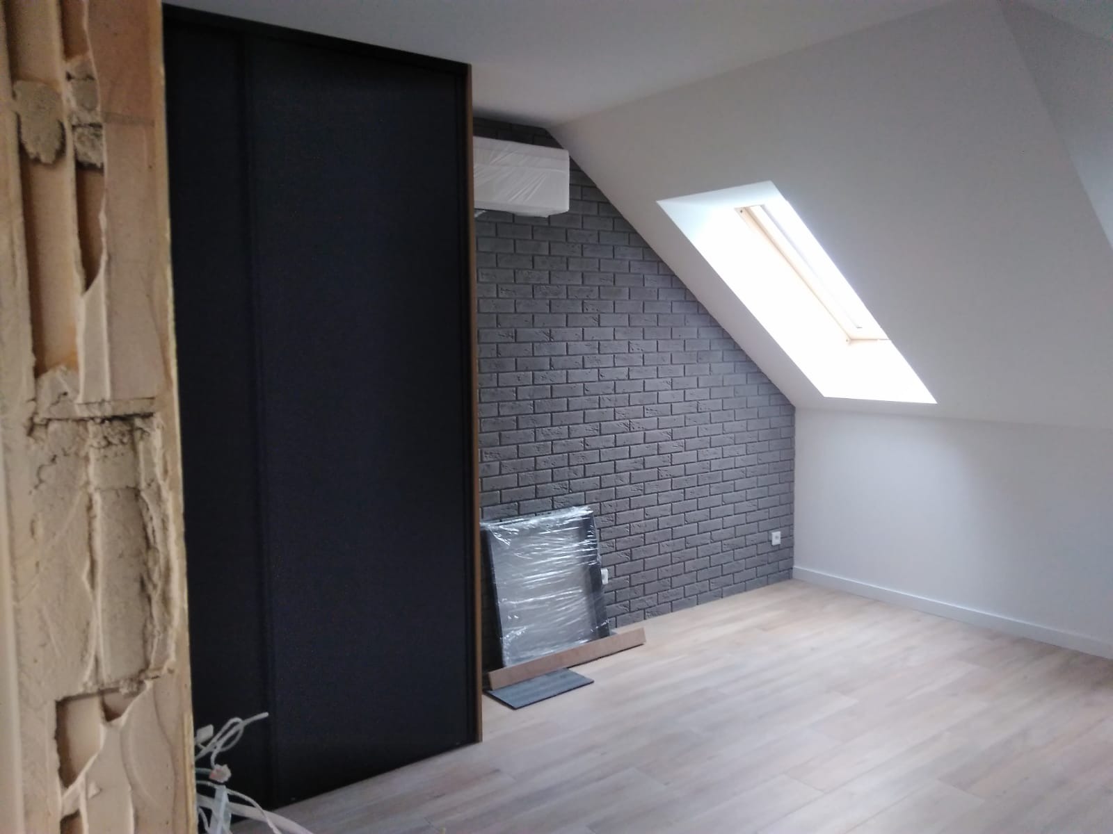 Wnętrze poddasza w trakcie remontu: fragment ściany z cegły dekoracyjnej, zamontowane drzwi przesuwne, surowe ściany z widocznym tynkiem oraz okno dachowe.