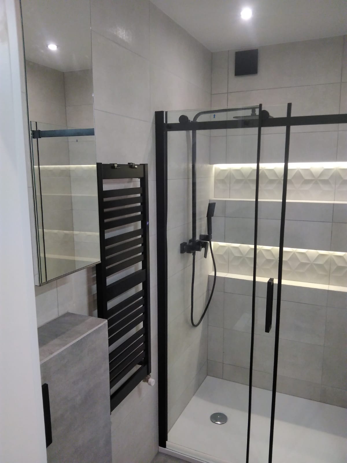 Nowoczesna łazienka z kabiną prysznicową w czarnej ramie, grzejnikiem drabinkowym w kolorze czarnym, umywalką z szarego betonu architektonicznego i podświetlanymi płytkami dekoracyjnymi na ścianie.