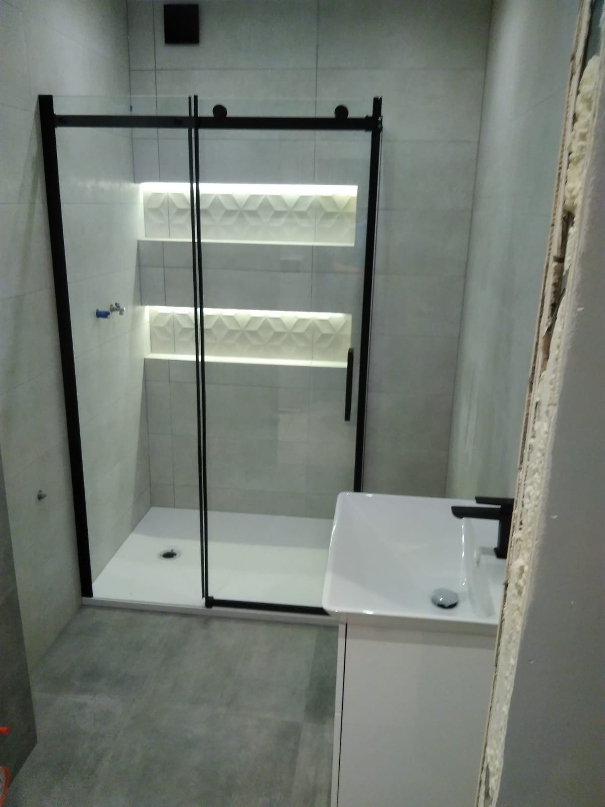 Nowoczesna łazienka z kabiną prysznicową z czarnymi ramami, umywalką na szafce i podświetlanymi wnękami w ścianie wyłożonej szarymi płytkami.