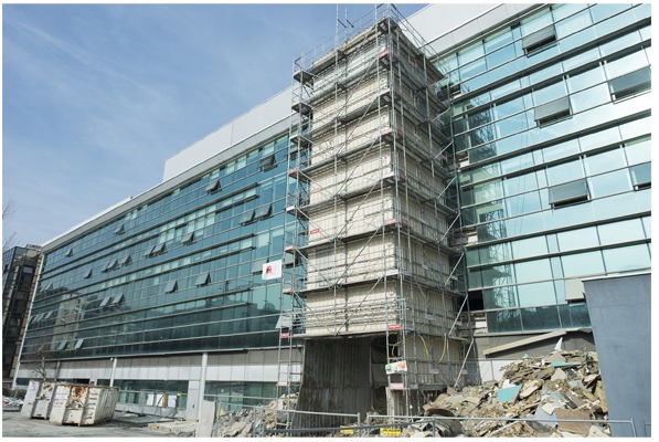 Fragment budynku biurowego w trakcie rozbiórki, osłonięty rusztowaniami, z widocznym stosem gruzu u podstawy i otwartymi oknami na pozostałej części elewacji.