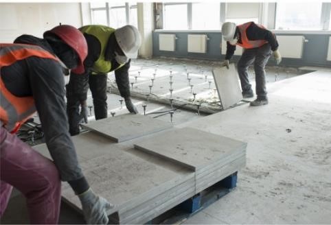 Pracownicy w MERU układają podniesioną podłogę na regulowanych wspornikach w remontowanym pomieszczeniu, widoczne płyty podłogowe na palecie i przygotowane podłoże.