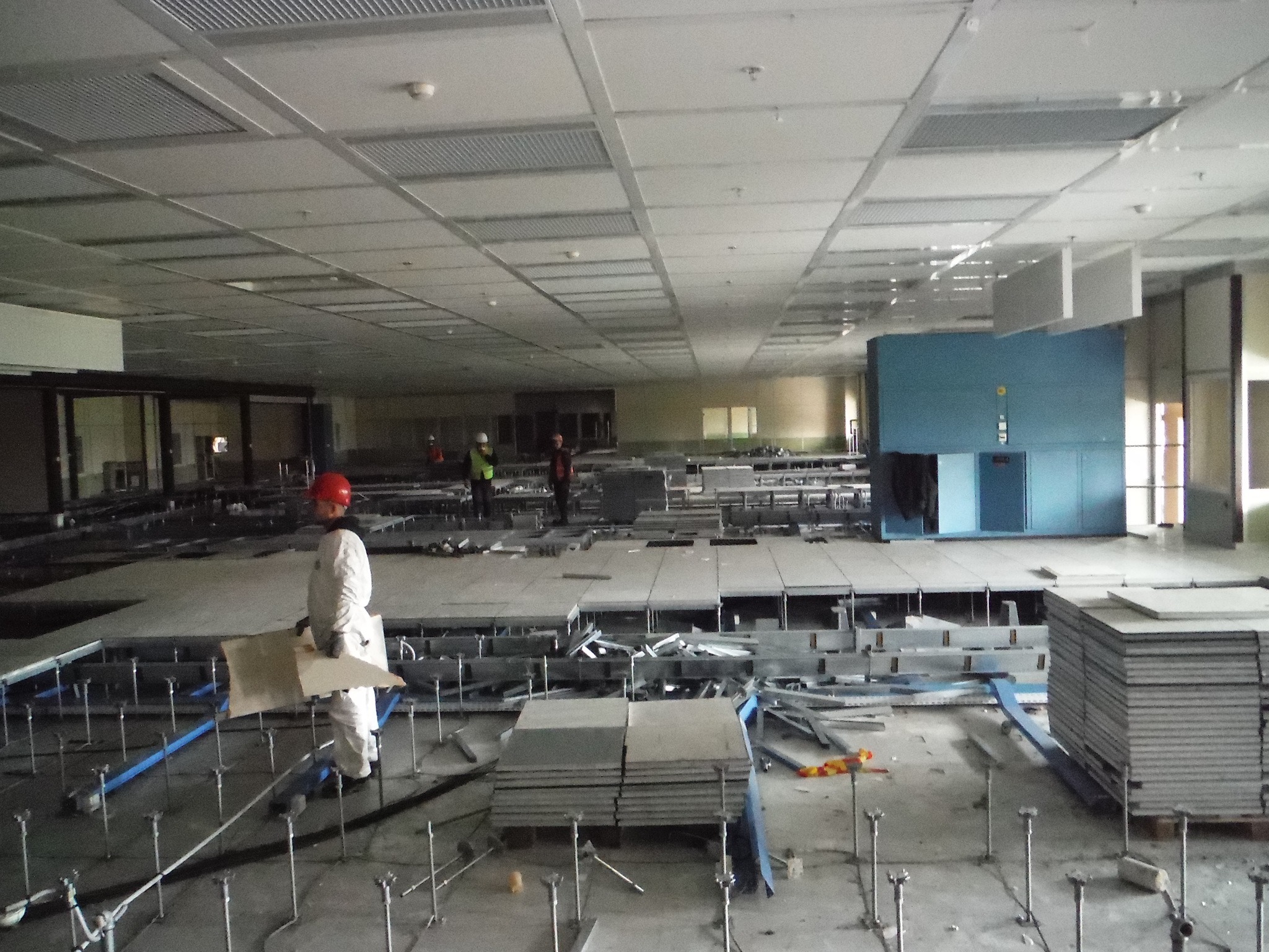 Przestronne wnętrze w trakcie prac demontażowych: pracownik w białym kombinezonie i czerwonym kasku trzyma płytę, w tle widoczne metalowe elementy konstrukcyjne i fragmenty podłogi podniesionej.