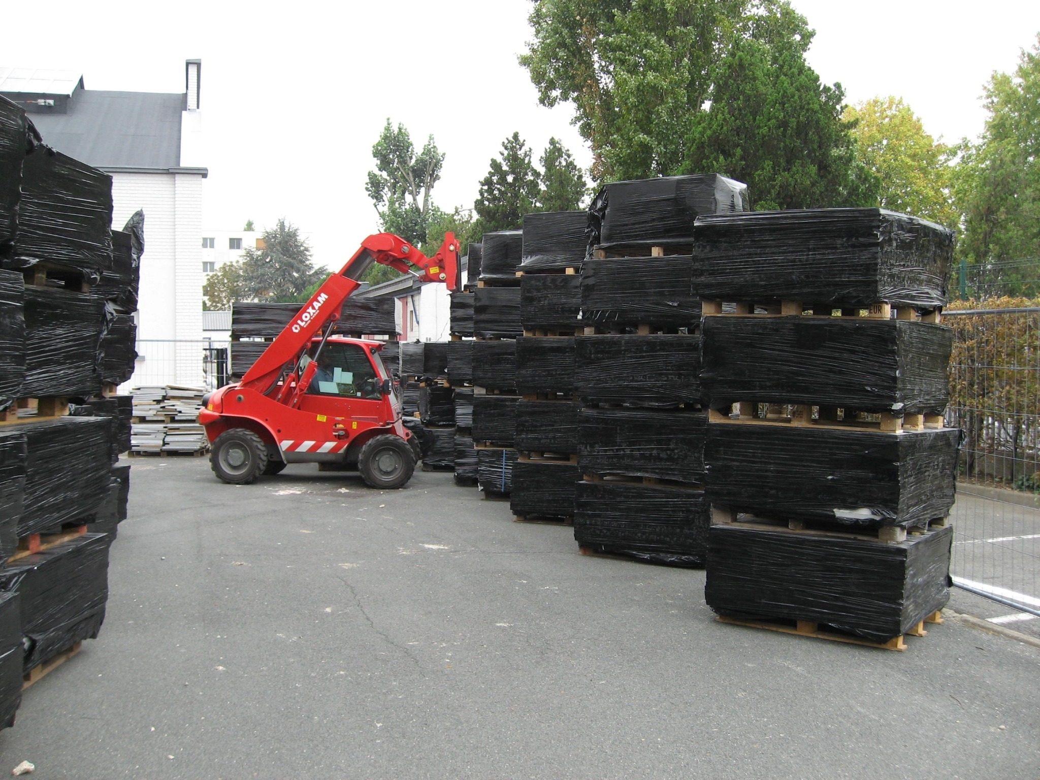 Czerwony wózek widłowy Oloxam manewruje w pobliżu palet z czarnymi płytami budowlanymi, przygotowując je do transportu lub składowania.