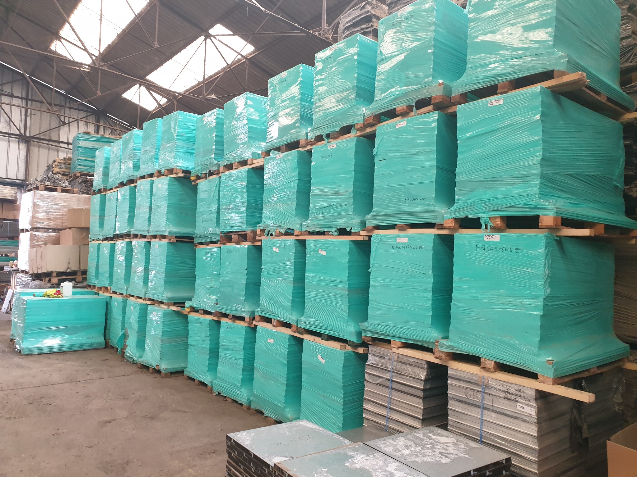 Stosy materiałów izolacyjnych, owiniętych w turkusową folię, ułożone na drewnianych paletach w magazynie z metalową konstrukcją dachu.