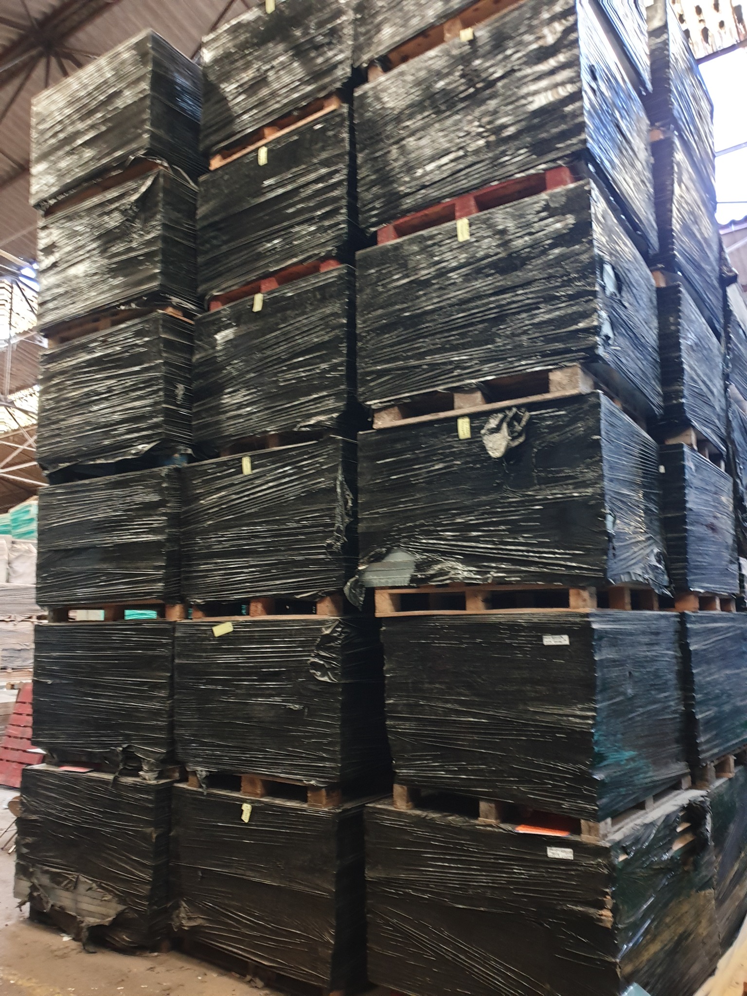 Stos palet z płytami zabezpieczonymi folią stretch, składowane w magazynie, widoczne etykiety na niektórych paczkach.