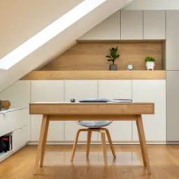Biurko z laptopem i notatnikiem w pokoju na poddaszu, z zabudową meblową w odcieniach bieli i szarości, drewniana podłoga i akcenty z jasnego drewna.