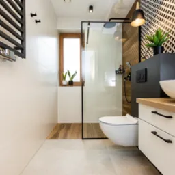 Nowoczesna łazienka z kabiną prysznicową typu walk-in, wiszącą toaletą, umywalką nablatową na szafce z jasnym blatem, grzejnikiem drabinkowym i oknem z roślinami doniczkowymi.