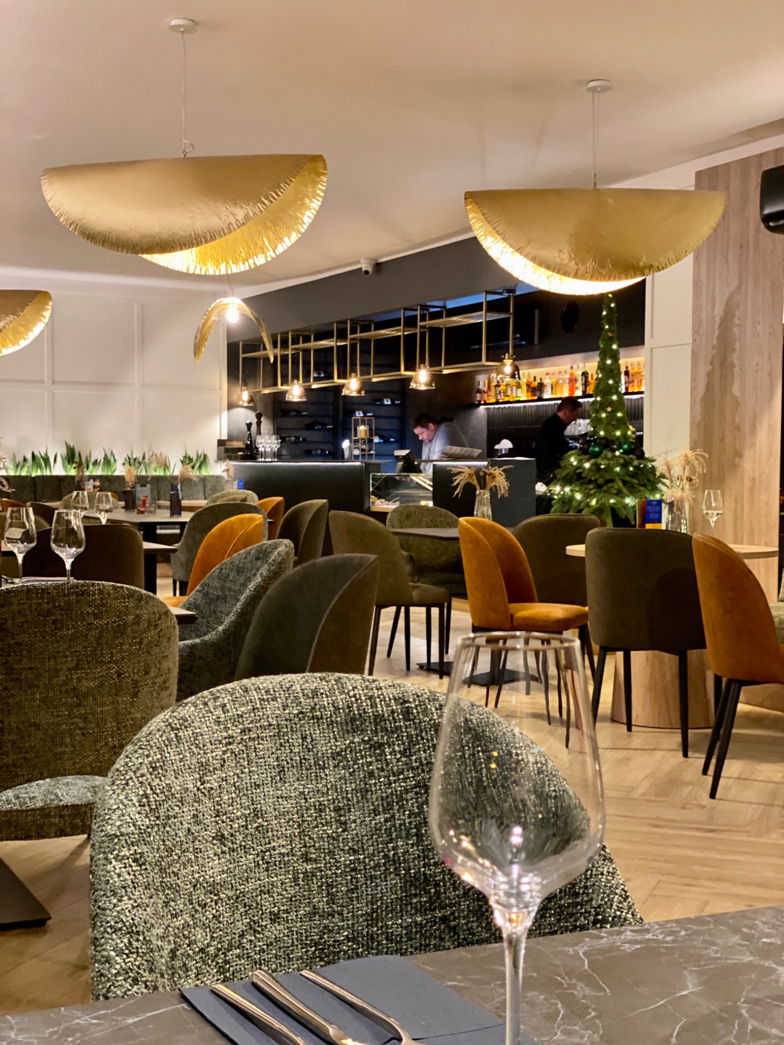 Wnętrze restauracji z oświetleniem w kształcie półksiężyca, stolik nakryty dla gości, w tle bar z pracującymi osobami i świąteczna choinka.