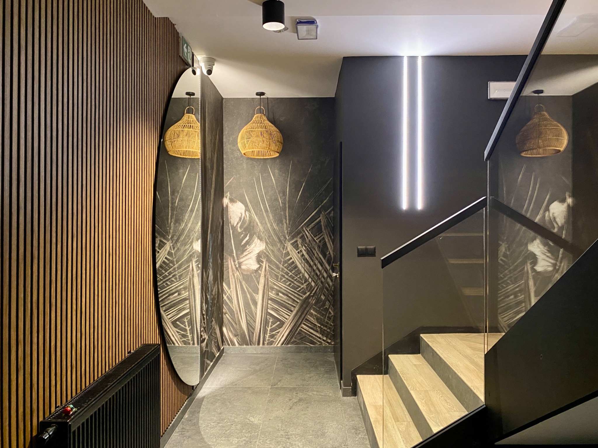 Nowoczesne wnętrze klatki schodowej z drewnianą boazerią, lustrem owalnym, schodami z drewnianymi stopniami i szklaną balustradą, oświetleniem LED i lampami wiszącymi w stylu boho.