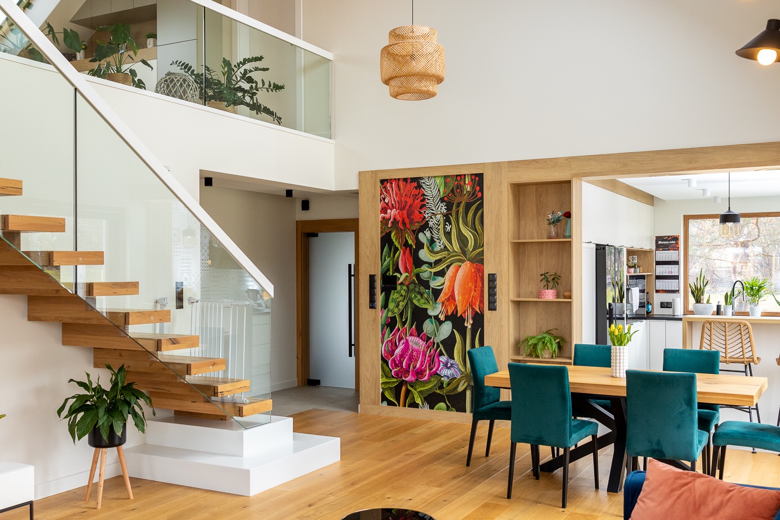Nowoczesne wnętrze domu z drewnianymi schodami ze szklaną balustradą, jadalnią z turkusowymi krzesłami i kuchnią w tle, akcentem kolorystycznym jest fototapeta z motywem kwiatowym na drzwiach.
