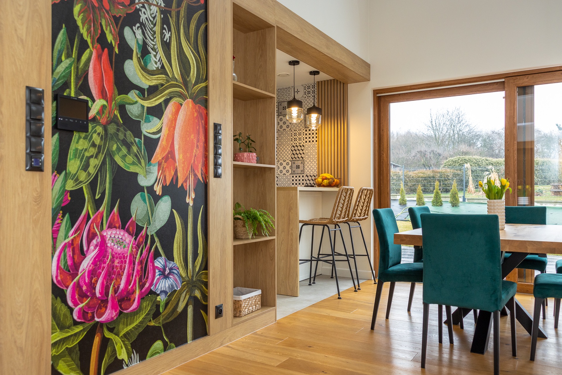 Nowoczesne wnętrze jadalni z drewnianą podłogą, ścianą z motywem kwiatowym, otwartą przestrzenią na kuchnię z hokerami i widokiem na ogród przez duże okno.
