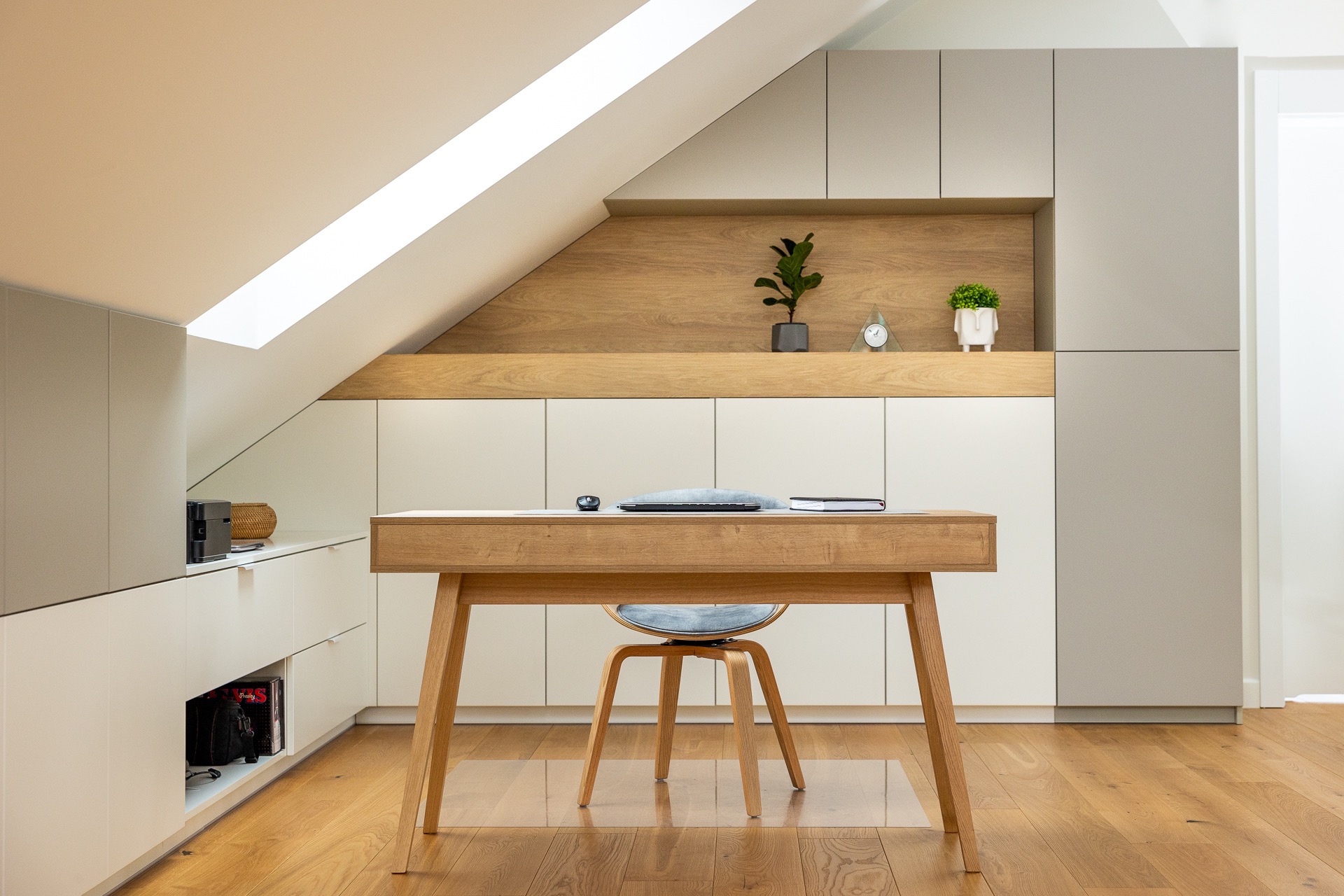 Biurko z laptopem i notatnikiem w pokoju na poddaszu, z zabudową meblową w odcieniach bieli i szarości, drewniana podłoga i akcenty z jasnego drewna.
