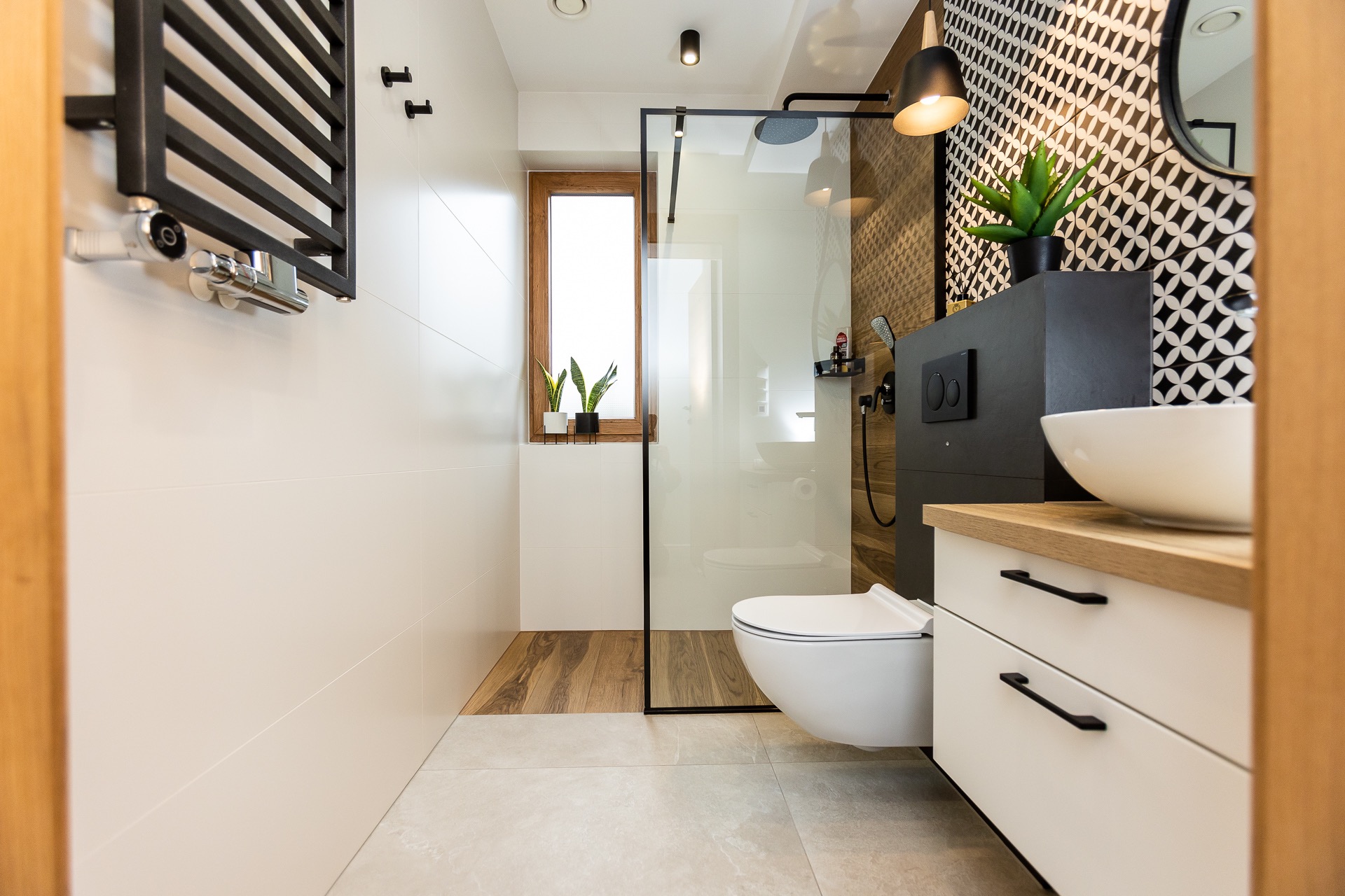 Nowoczesna łazienka z kabiną prysznicową typu walk-in, wiszącą toaletą, umywalką nablatową na szafce z jasnym blatem, grzejnikiem drabinkowym i oknem z roślinami doniczkowymi.