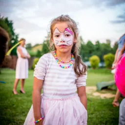 Dziewczynka z pomalowaną twarzą w motywie kota, ubrana w różową sukienkę i kolorową biżuterię, pozująca na tle zielonego trawnika podczas imprezy plenerowej.