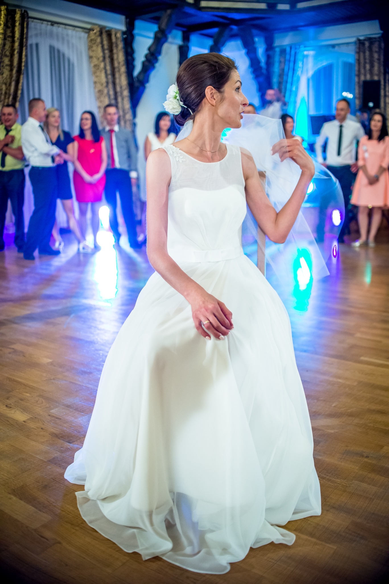 Panna młoda w białej sukni siedzi na krześle podczas wesela w Rzeszowie, z kwiatami we włosach i delikatnym welonem, otoczona gośćmi i dekoracjami sali.