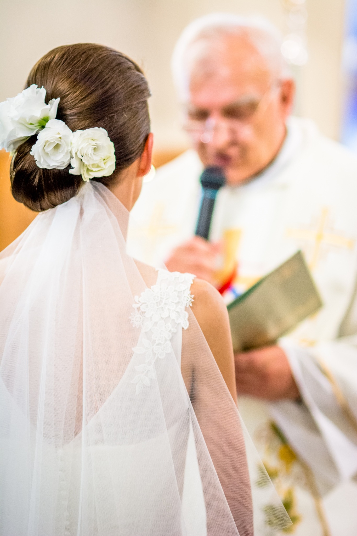 Panna młoda w białej sukni i welonie z kwiatami we włosach, podczas ceremonii ślubnej w kościele, ksiądz z mikrofonem w tle.