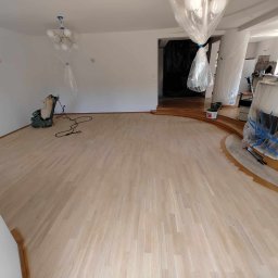 RENO-PAR VADYM SHPITSA - Duży, jasny salon z parkietem po wstępnym szlifowaniu, zabezpieczone folią meble i elementy wystroju, widoczna maszyna do szlifowania parkietu w rogu.