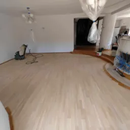Duży, jasny salon z parkietem po wstępnym szlifowaniu, zabezpieczone folią meble i elementy wystroju, widoczna maszyna do szlifowania parkietu w rogu.