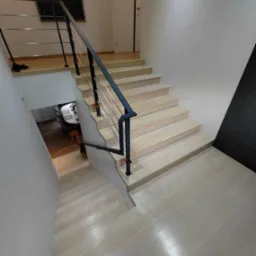 Nowoczesne, jasne drewniane schody z czarną metalową balustradą, widok z góry na dół, z widocznym fragmentem jadalni na dole oraz obrazem na ścianie.