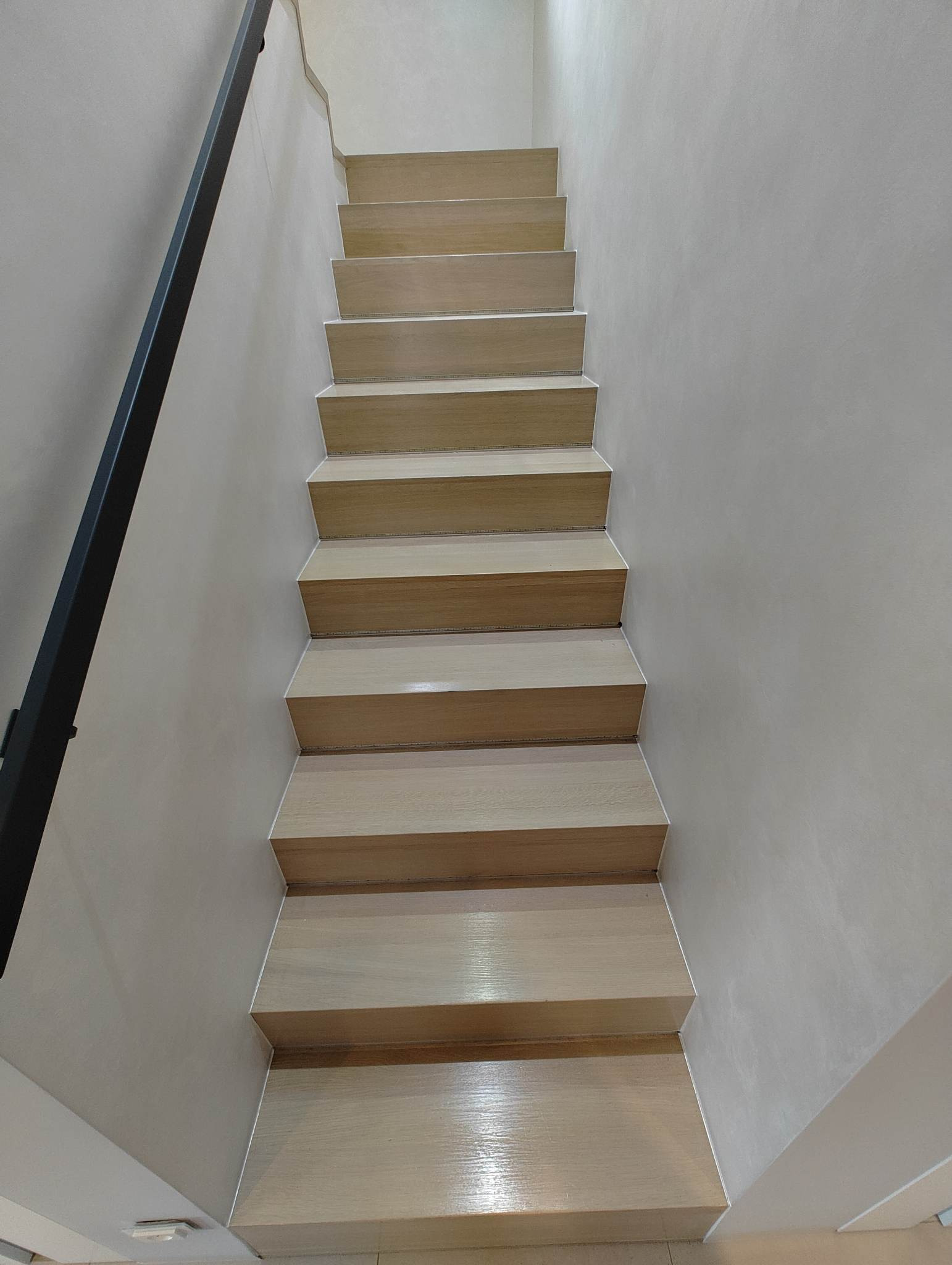 Jasne drewniane schody z czarną metalową balustradą, widok z dołu, ściany w odcieniach beżu.