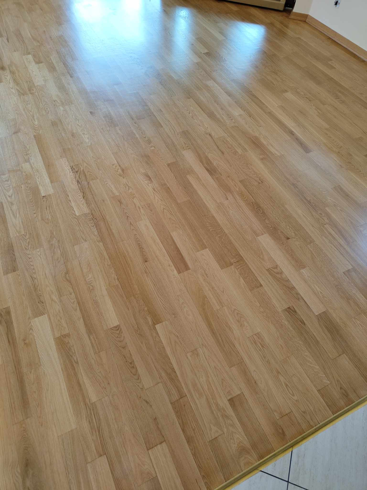 Świeżo ułożona, lakierowana podłoga panelowa w odcieniach jasnego dębu, z widocznymi refleksami światła i fragmentem białej listwy przypodłogowej z szarymi fugami.
