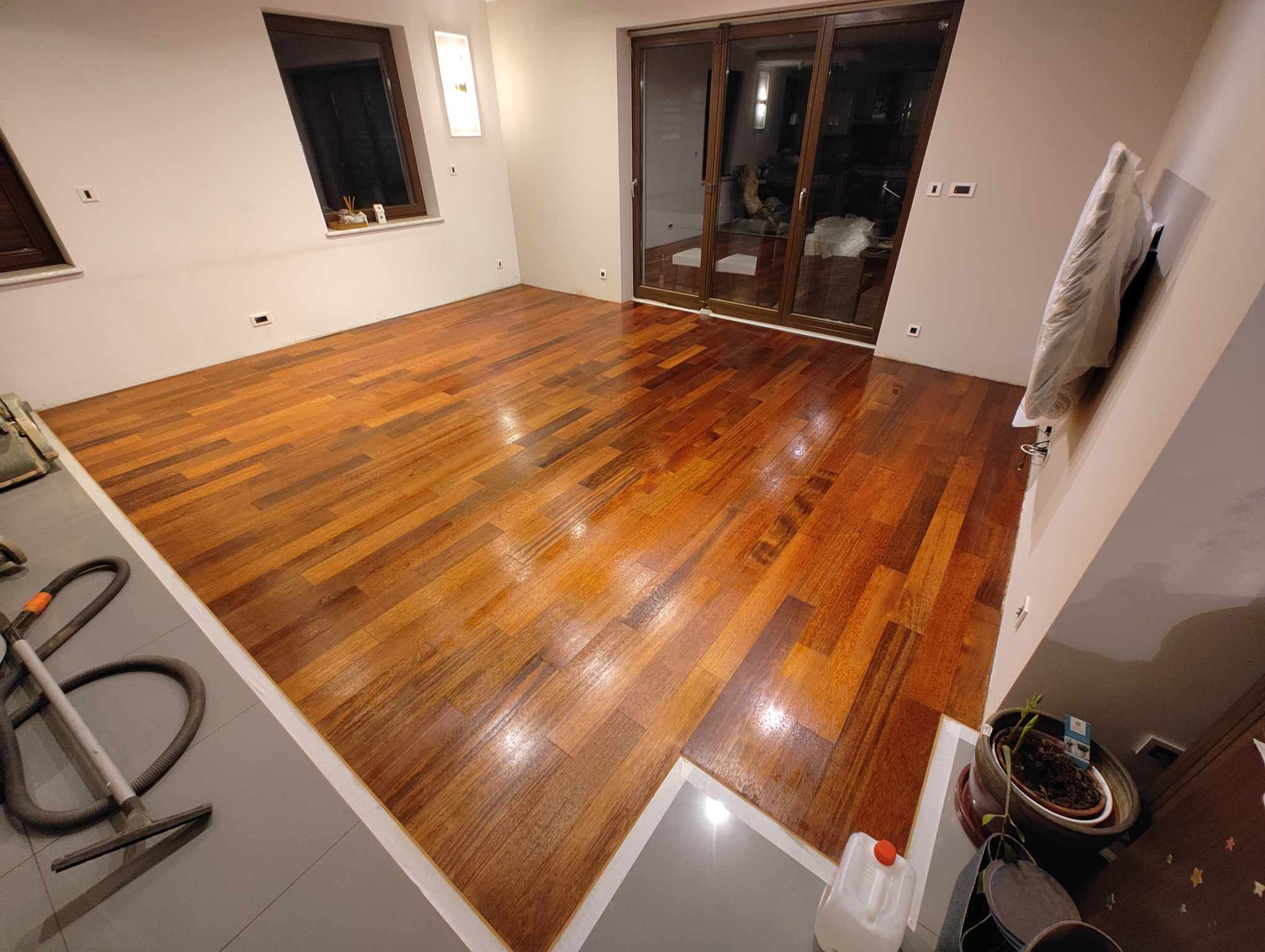 Świeżo polakierowana podłoga z egzotycznego drewna w przestronnym salonie, widoczne ślady po lakierowaniu i odkurzacz przemysłowy w rogu pomieszczenia.
