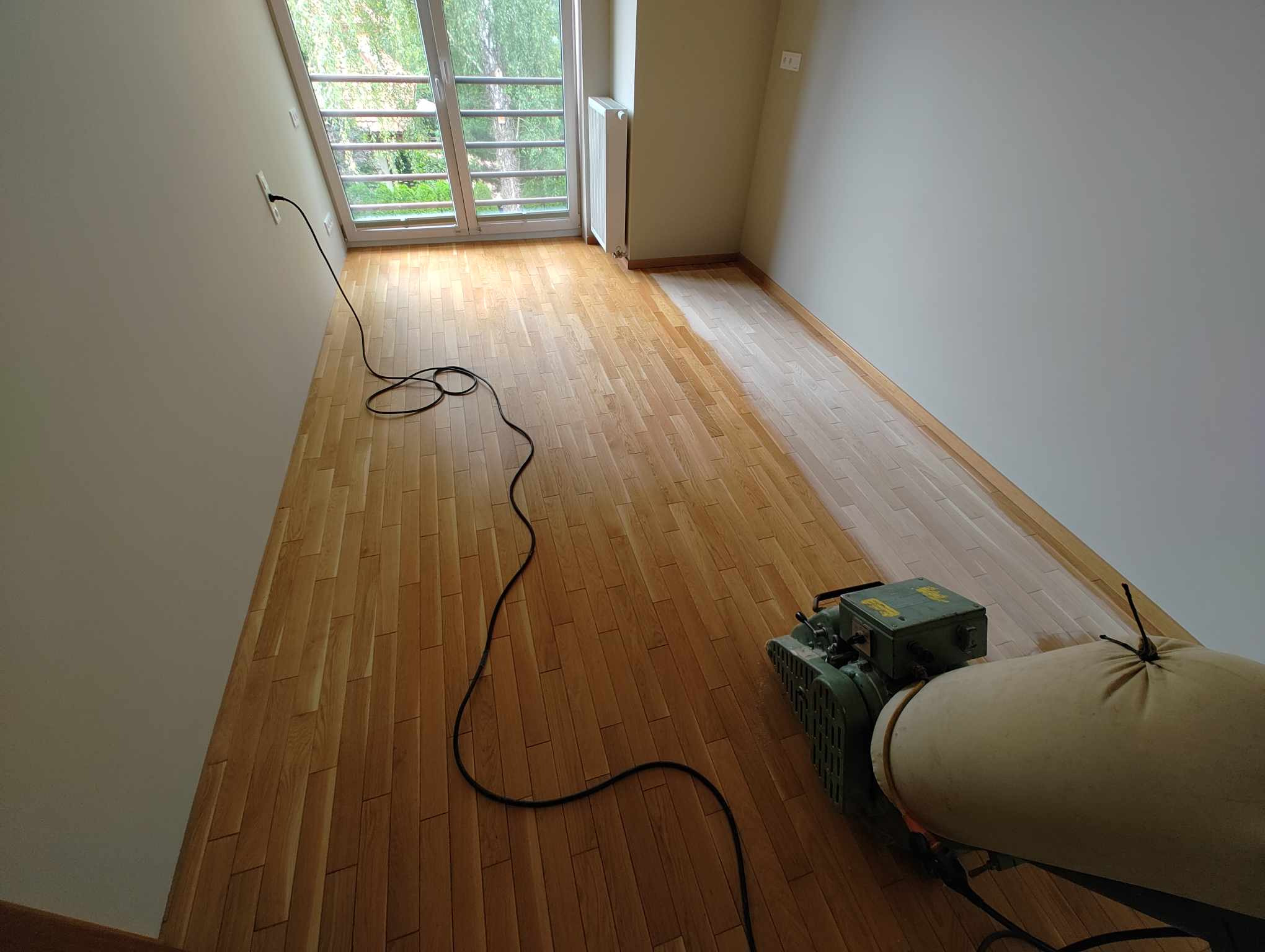Pokój z drewnianą podłogą w trakcie renowacji, część podłogi po obróbce, widoczna maszyna do szlifowania parkietu i kabel zasilający.