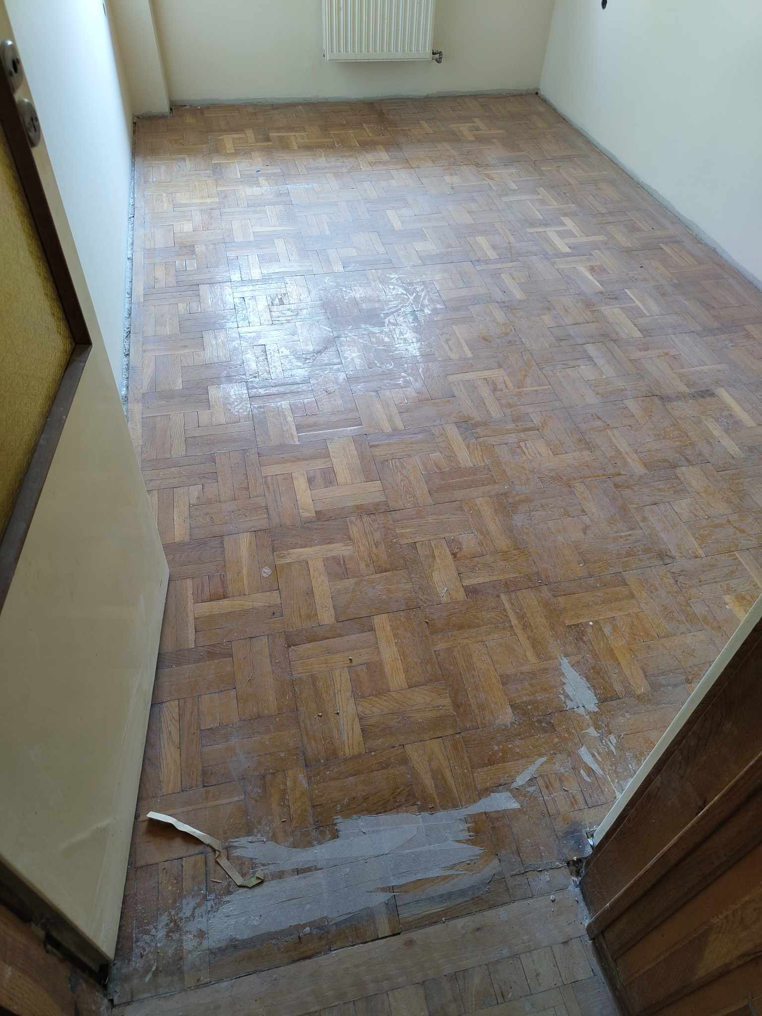 Fragment pokoju z parkietem ułożonym w jodełkę, widoczne ślady szpachli i niedoskonałości wykończenia, otwarte drzwi z lewej strony kadru.