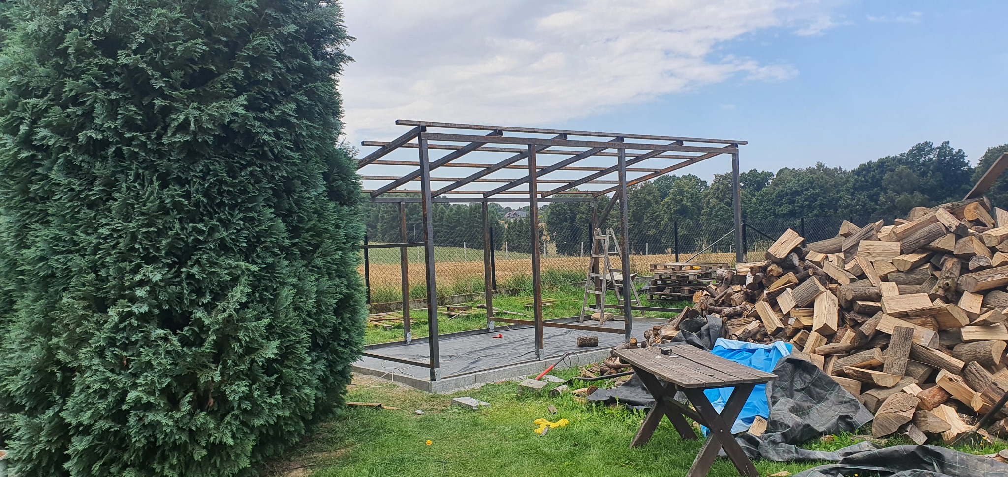 Konstrukcja altany ogrodowej w trakcie budowy, widoczny szkielet z metalowych profili na betonowej wylewce, obok stos drewna opałowego i stolik.
