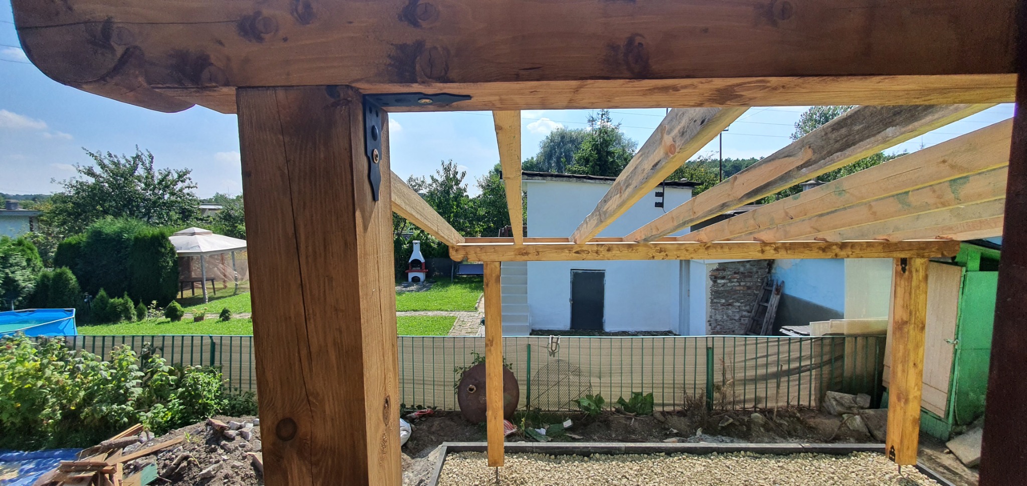 Drewniana konstrukcja altany w trakcie budowy, widok z wnętrza, z widocznymi elementami mocującymi, w tle ogród z altaną, basenem i grillem.