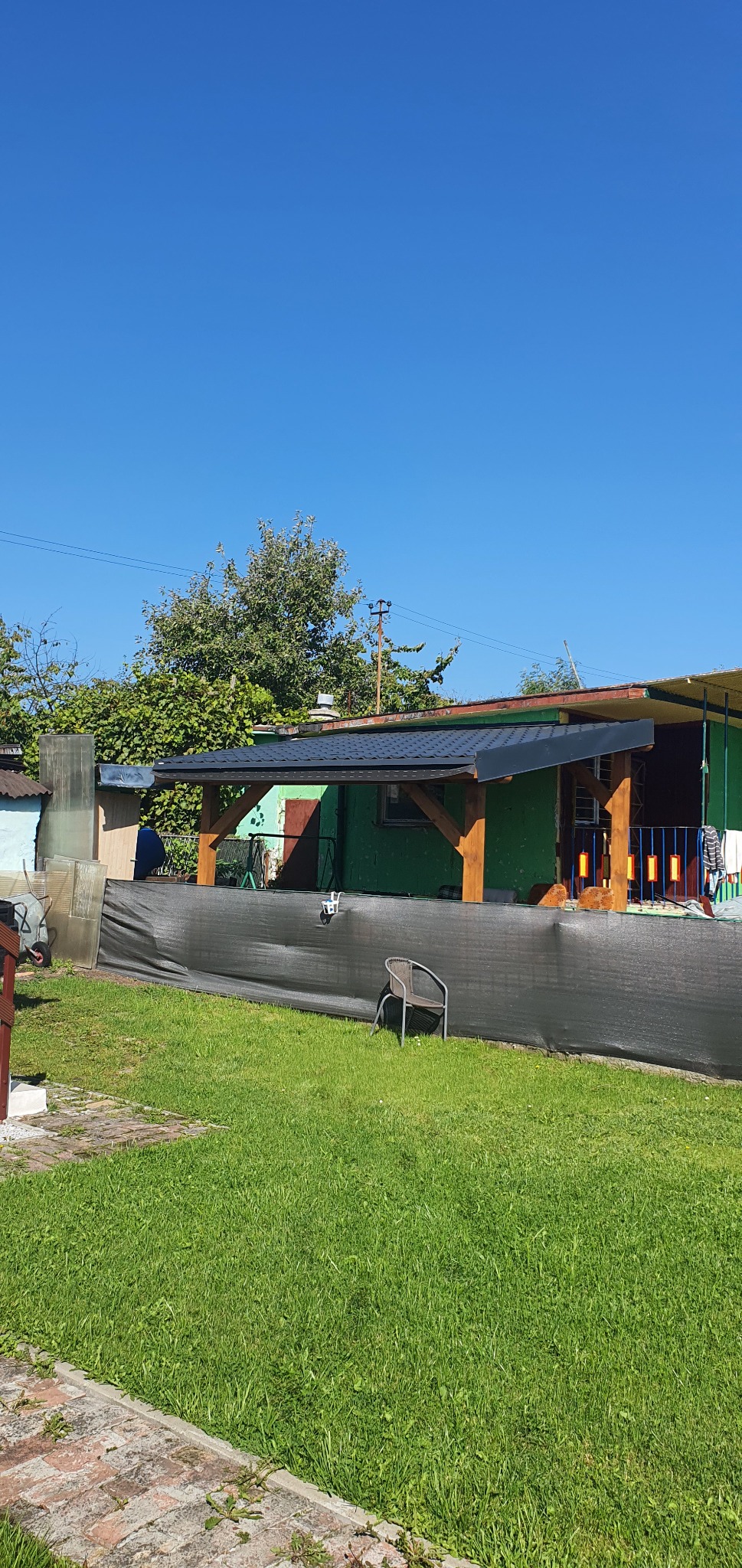 Zadaszenie tarasu z drewnianą konstrukcją i czarnym dachem, widoczne z trawnika, z krzesłem ogrodowym w tle.