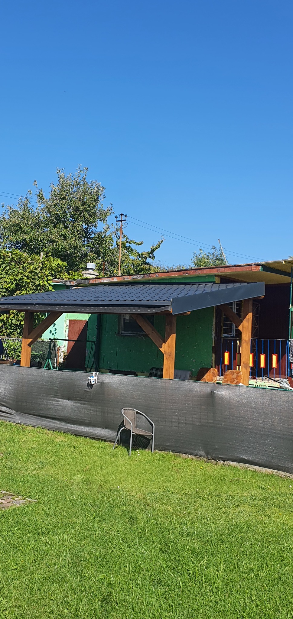 Zadaszenie tarasu z drewnianą konstrukcją i czarną dachówką, częściowo osłonięte czarną siatką cieniującą, z krzesłem na trawniku przed nim.