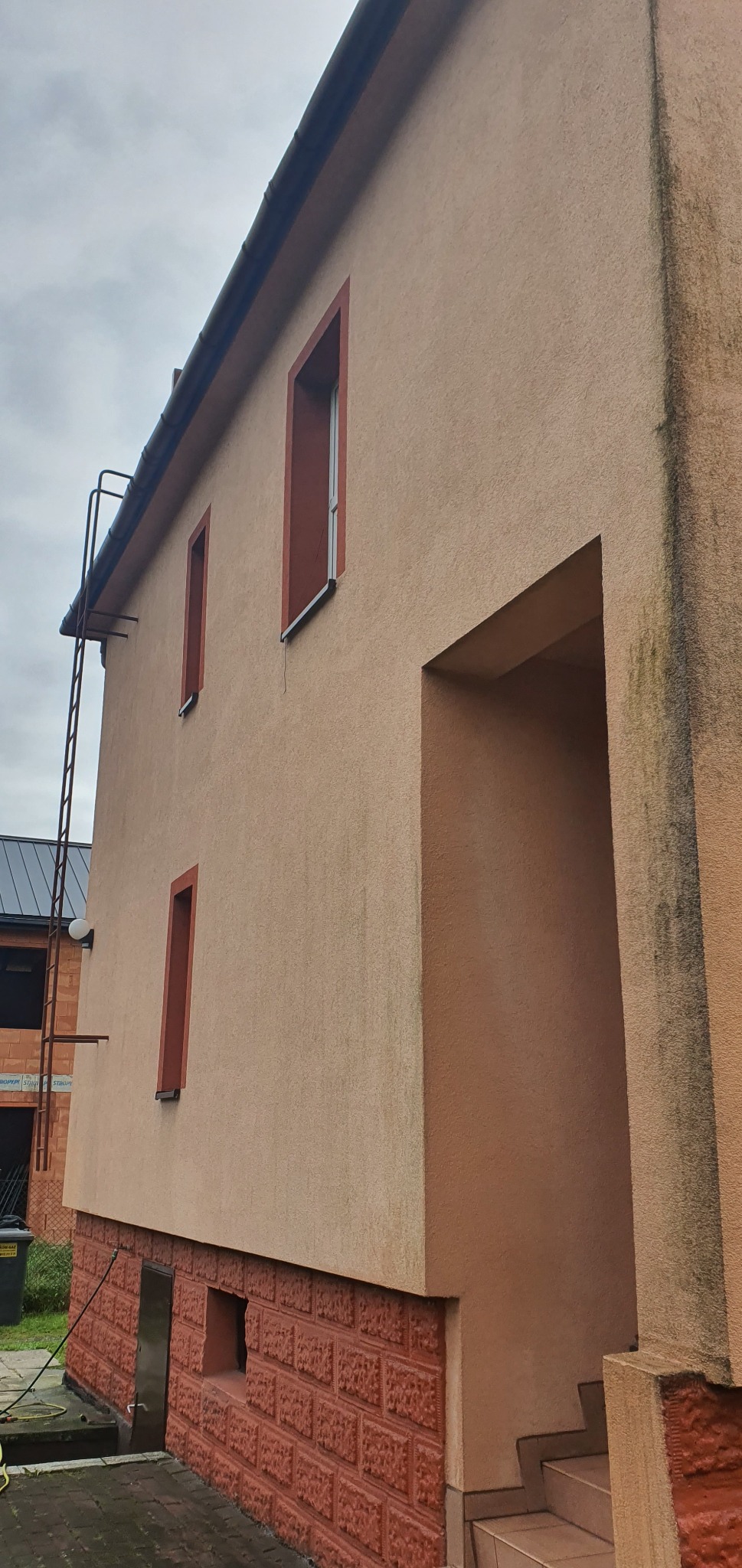 Fragment elewacji budynku z oknami w bordowych ramach, widoczna drabina na dachu i wejście do budynku z widocznymi schodami, dolna część elewacji wykończona czerwoną cegłą.