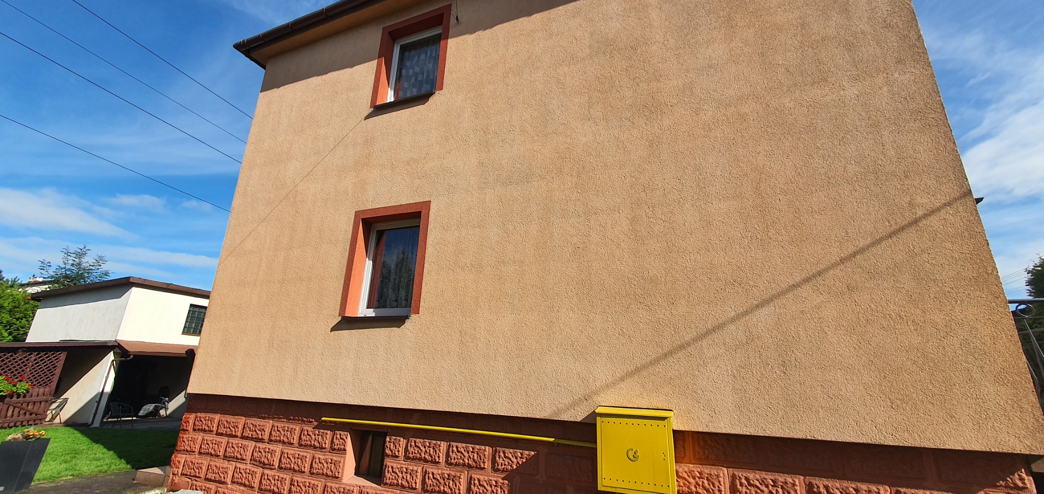 Fragment elewacji budynku z oknami w pomarańczowych ramach i żółtą skrzynką gazową, widoczny fragment ceglanego cokołu.