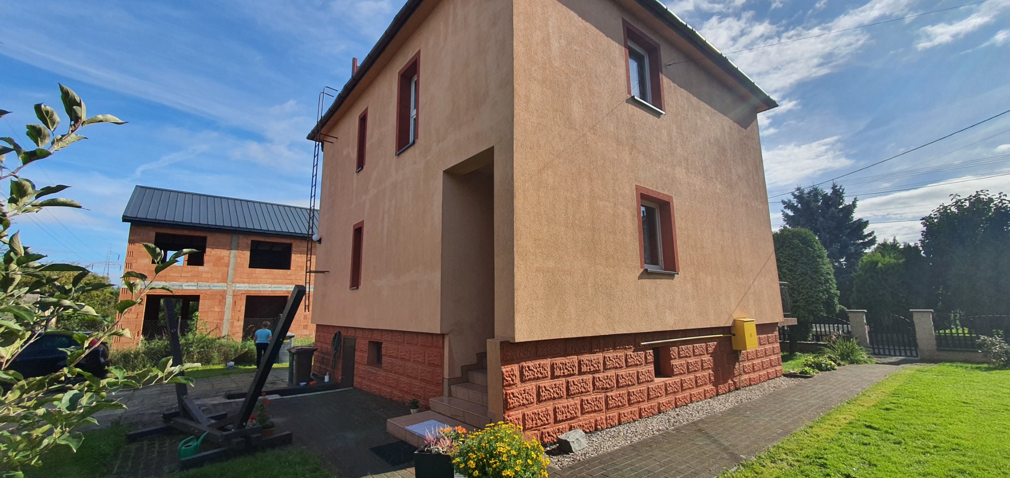 Dwupiętrowy dom z tynkowaną elewacją w odcieniach beżu i brązu, z czerwonymi obramowaniami okien i dekoracyjnym cokołem z cegły, na tle błękitnego nieba i zieleni ogrodu.