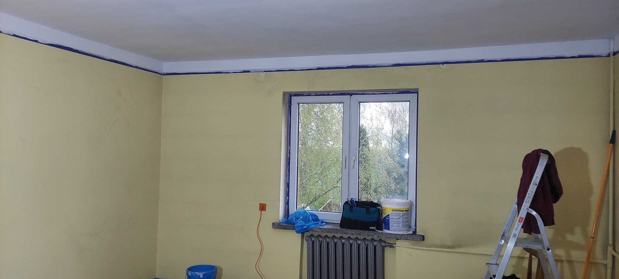 Pokój w trakcie przygotowania do malowania: ściany pomalowane na żółto, okno i listwy zabezpieczone taśmą malarską, widoczna drabina, wiadro z farbą i narzędzia.