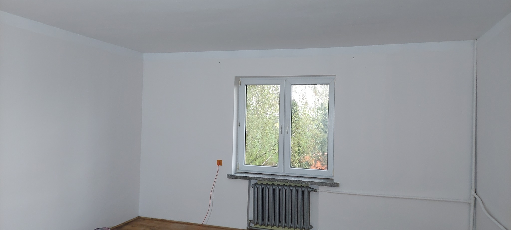 Pomalowane na biało wnętrze pokoju z widokiem na zieleń za oknem, szary grzejnik pod parapetem i wystający kabel elektryczny z pomarańczowej puszki na ścianie.