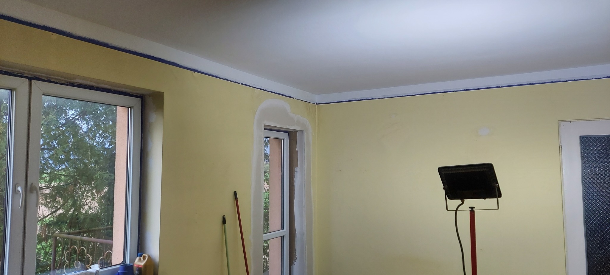 Żółta ściana w trakcie przygotowania do malowania, z widocznymi fragmentami szpachli wokół okien, zabezpieczona taśmą malarską przy suficie, oświetlona lampą budowlaną.