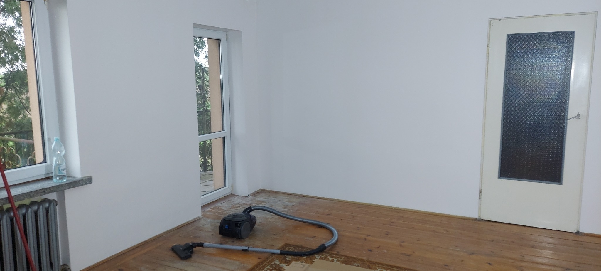Pokój po remoncie: odkurzacz na drewnianej podłodze, świeżo pomalowane białe ściany, okno z widokiem na zieleń i butelka wody na marmurowym parapecie.