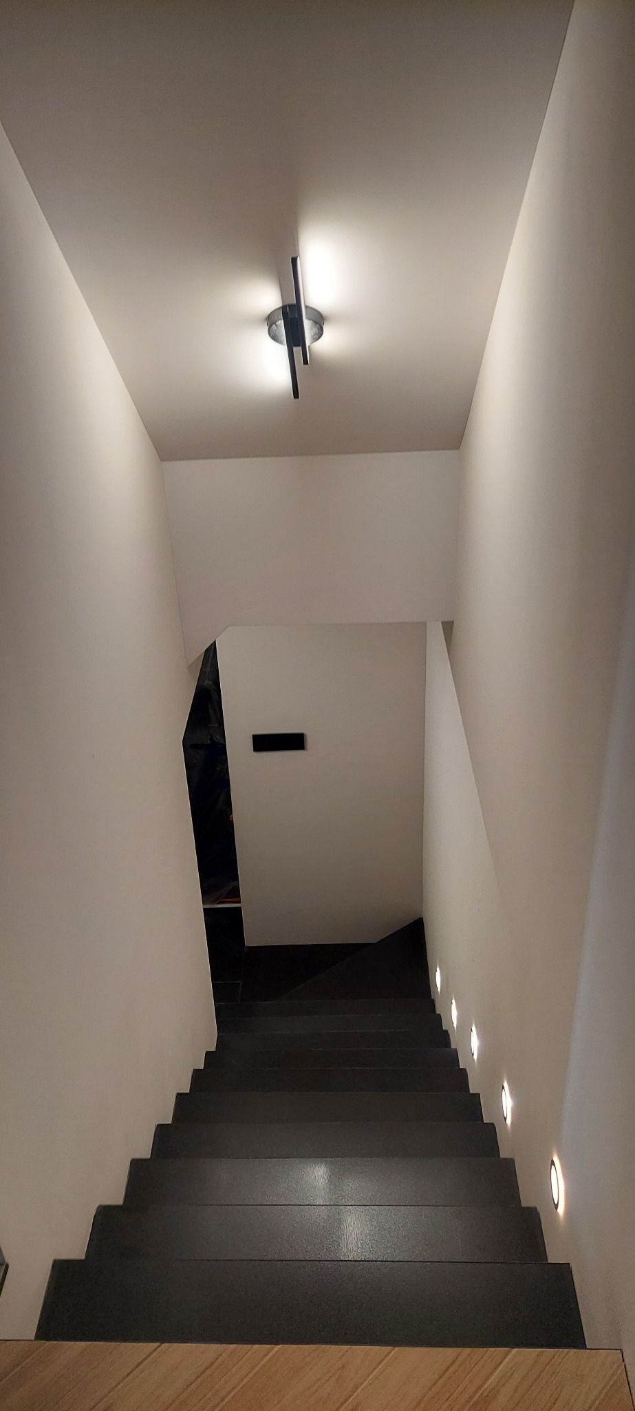 Nowoczesne schody z ciemnego kamienia z podświetleniem punktowym w ścianie i lampą sufitową o geometrycznym kształcie.