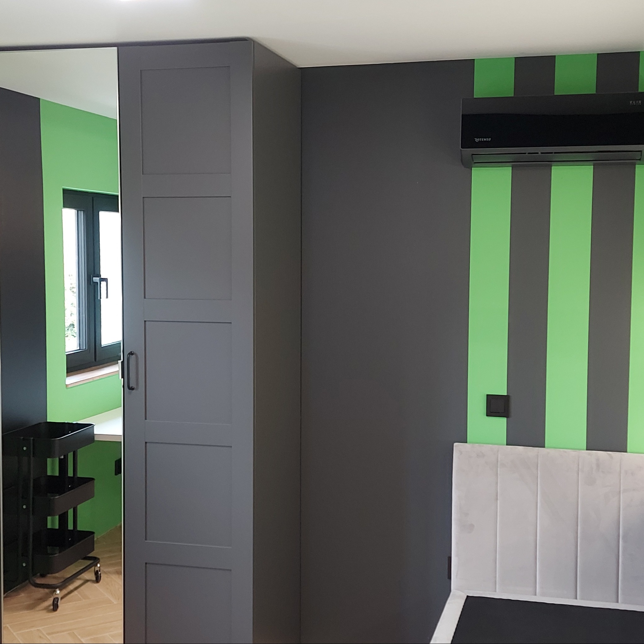 Szara szafa z lustrzanymi drzwiami przesuwnymi, odbijająca fragment pokoju z zielonymi ścianami, czarnym regałem na kółkach i oknem; ściana w szaro-zielone pasy z czarnym klimatyzatorem...