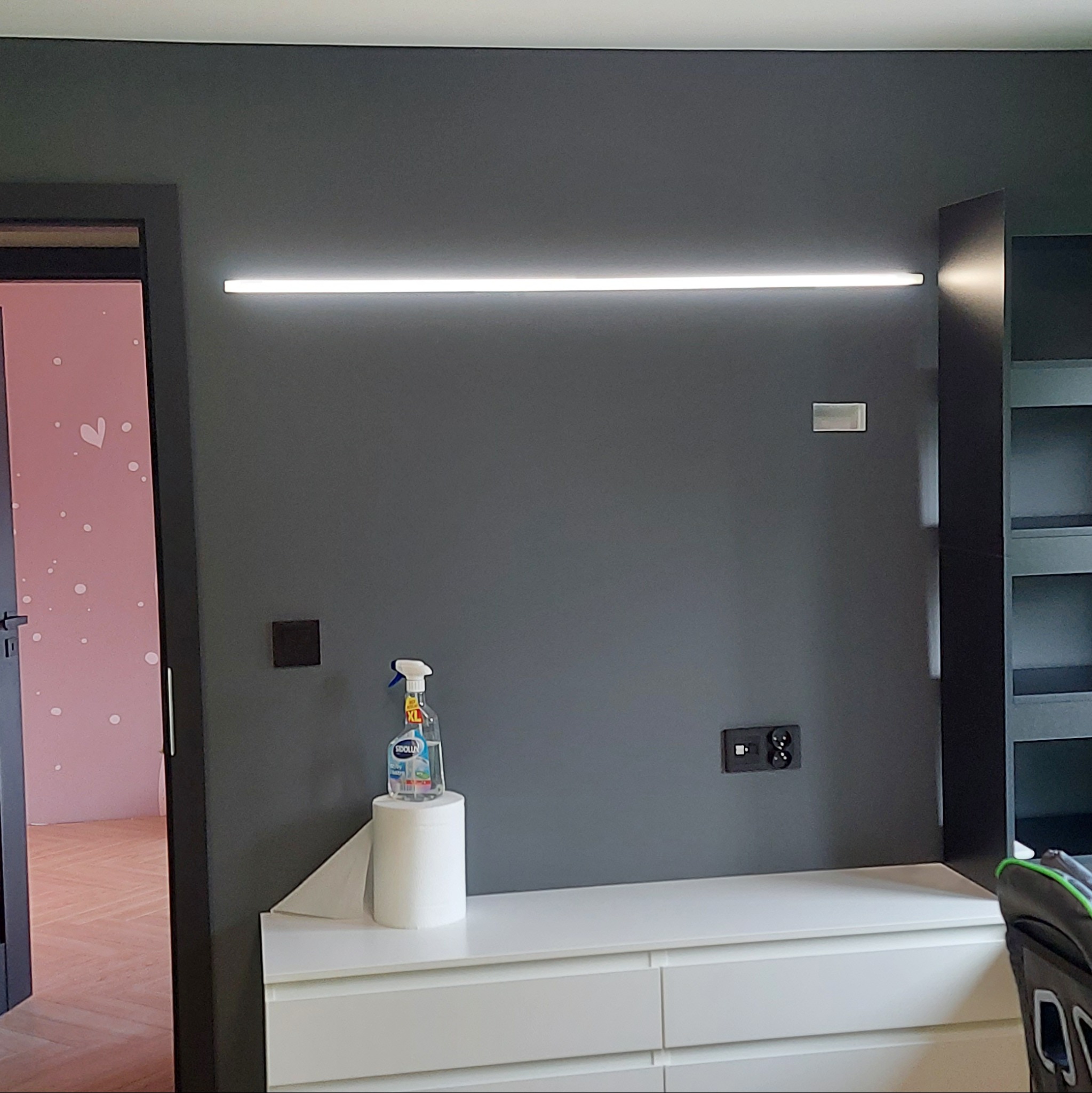 Szara ściana z podłużnym oświetleniem LED, gniazdko elektryczne, w tle różowy pokój z dekoracjami w kształcie serc widoczny przez otwarte drzwi, na białej komodzie stoi środek czyszczący i rolka...