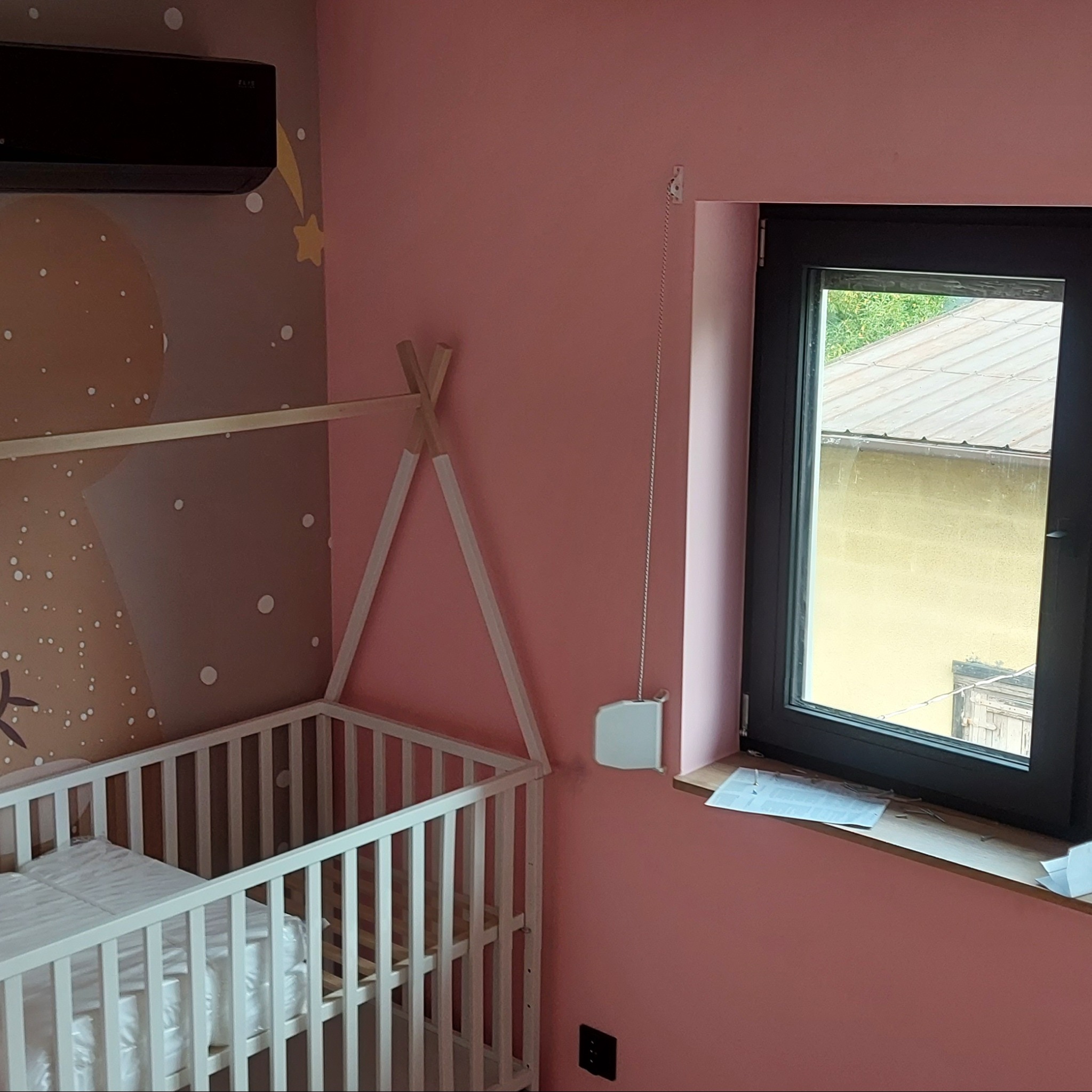 Pokój dziecięcy z różowymi ścianami, białym łóżeczkiem z baldachimem w stylu tipi, czarną klimatyzacją i oknem z widokiem na zewnątrz.