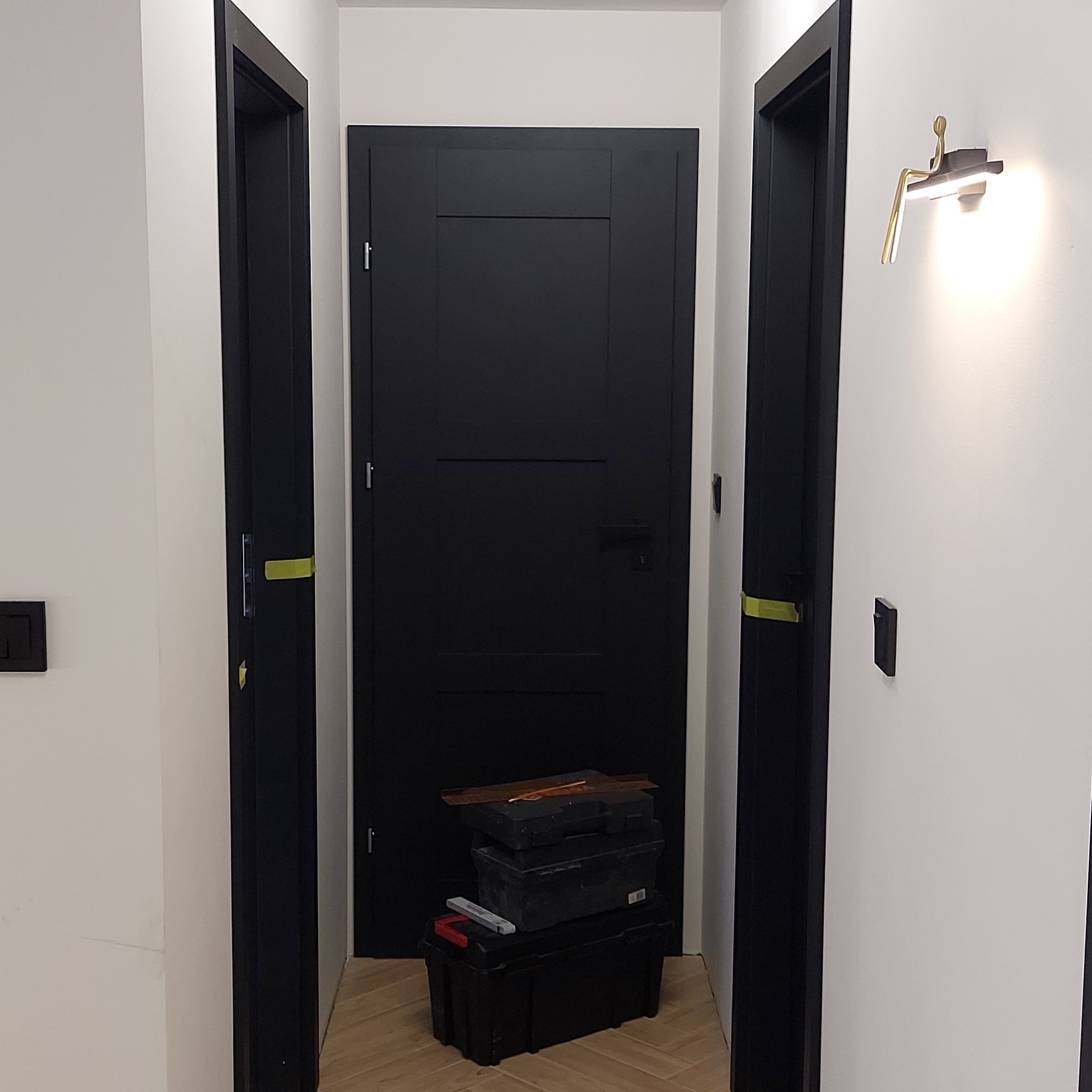 Montaż czarnych, minimalistycznych drzwi wewnętrznych; widoczne narzędzia i skrzynki monterskie, nowoczesna lampa ścienna w kształcie ludzika.