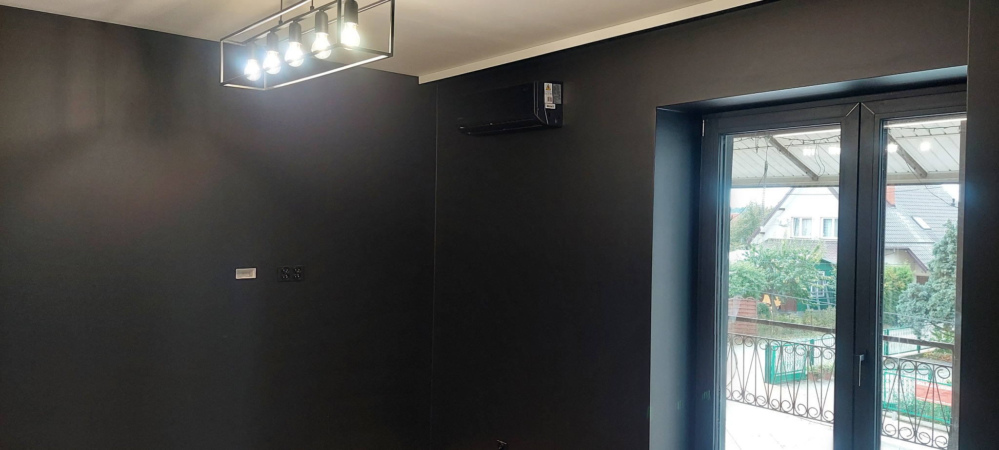 Ciemnoszary pokój po remoncie z czarnym klimatyzatorem, nowoczesną lampą sufitową i oknem balkonowym z widokiem na zewnątrz.