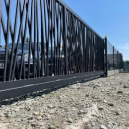 Czarna brama wjazdowa z pionowymi, nieregularnymi prętami, widok z dołu na żwirową nawierzchnię, w tle zaparkowany ciemny samochód i błękitne niebo.