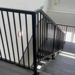 Klatka schodowa z czarną, metalową balustradą z pionowymi prętami; widoczne stopnie z antypoślizgowymi paskami i lampa ścienna.