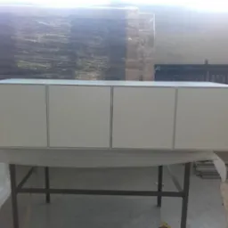 Biała szafka wisząca z czterema drzwiczkami, umieszczona na metalowym stole w warsztacie, z widocznymi materiałami budowlanymi w tle.