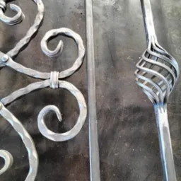 Elementy dekoracyjne z metalu, prawdopodobnie fragment balustrady lub bramy, z widocznymi spawami i spiralnym zdobieniem, leżące na roboczym, metalowym stole.
